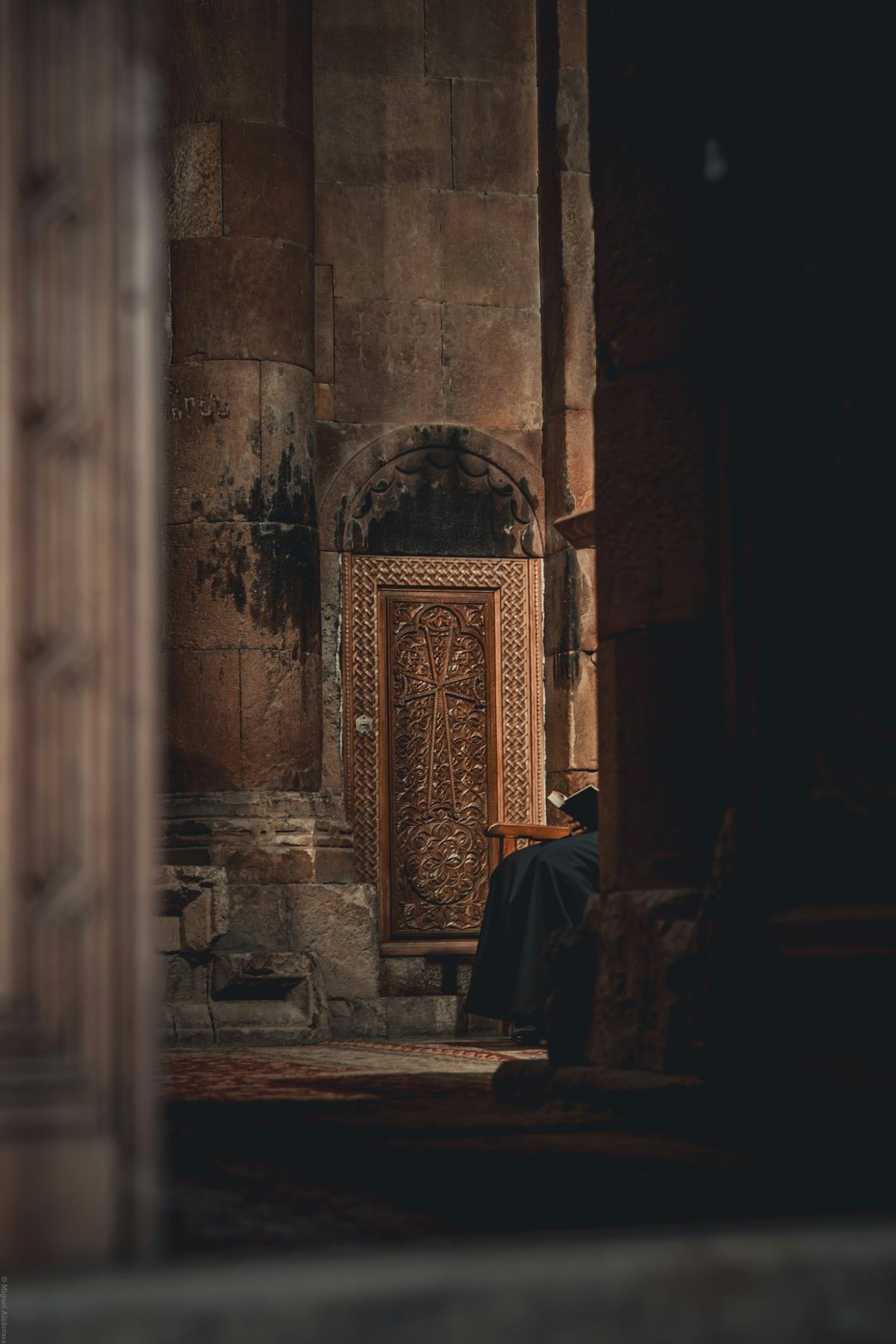 A monk in a black robe sits reading beside a carved wooden door in an old stone church, seen through a dark doorway.